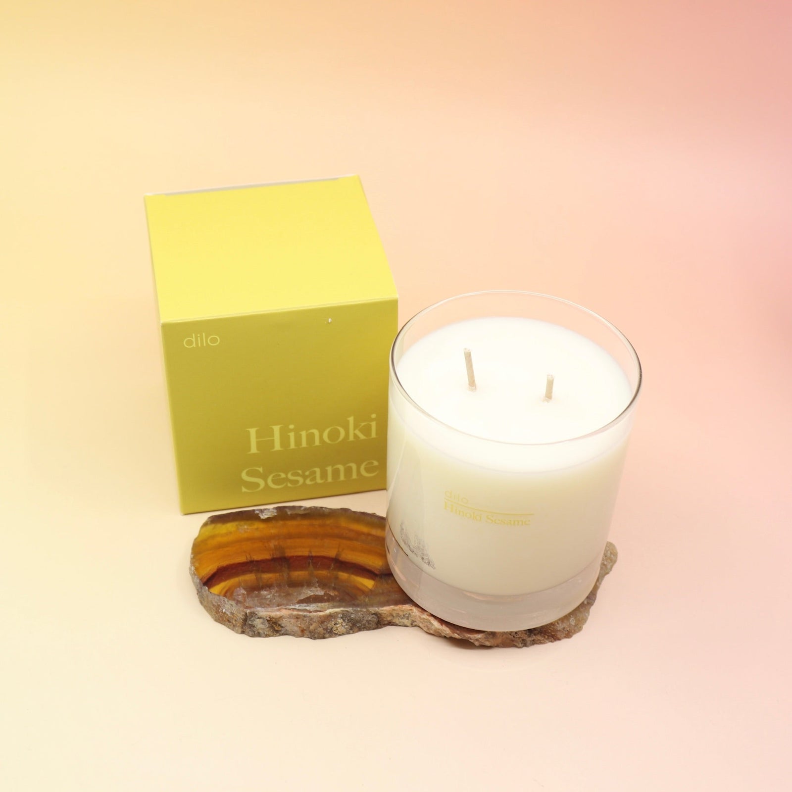 The Hinoki Sesame Candle on an agate slice next to its box all on a pink and yellow background.