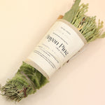 The Piñon Pine Sacred Smoke Cleansing Stick with Quartz Crystal on a pink and yellow background.