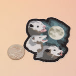 The Three Possum Moon Vinyl Sticker next to a quarter on a pink and yellow background.