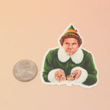 The Buddy the Elf Eating Spaghetti Sticker next to a quarter on a pink and yellow background.