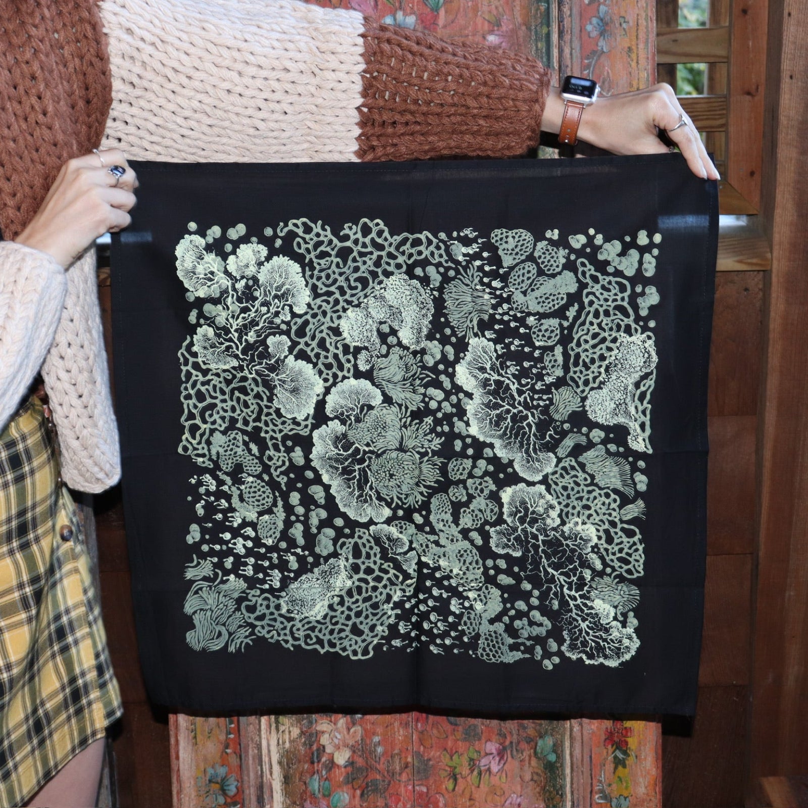 Someone holding up the Black Slime Mold Cotton Bandana in front of a wooden background.