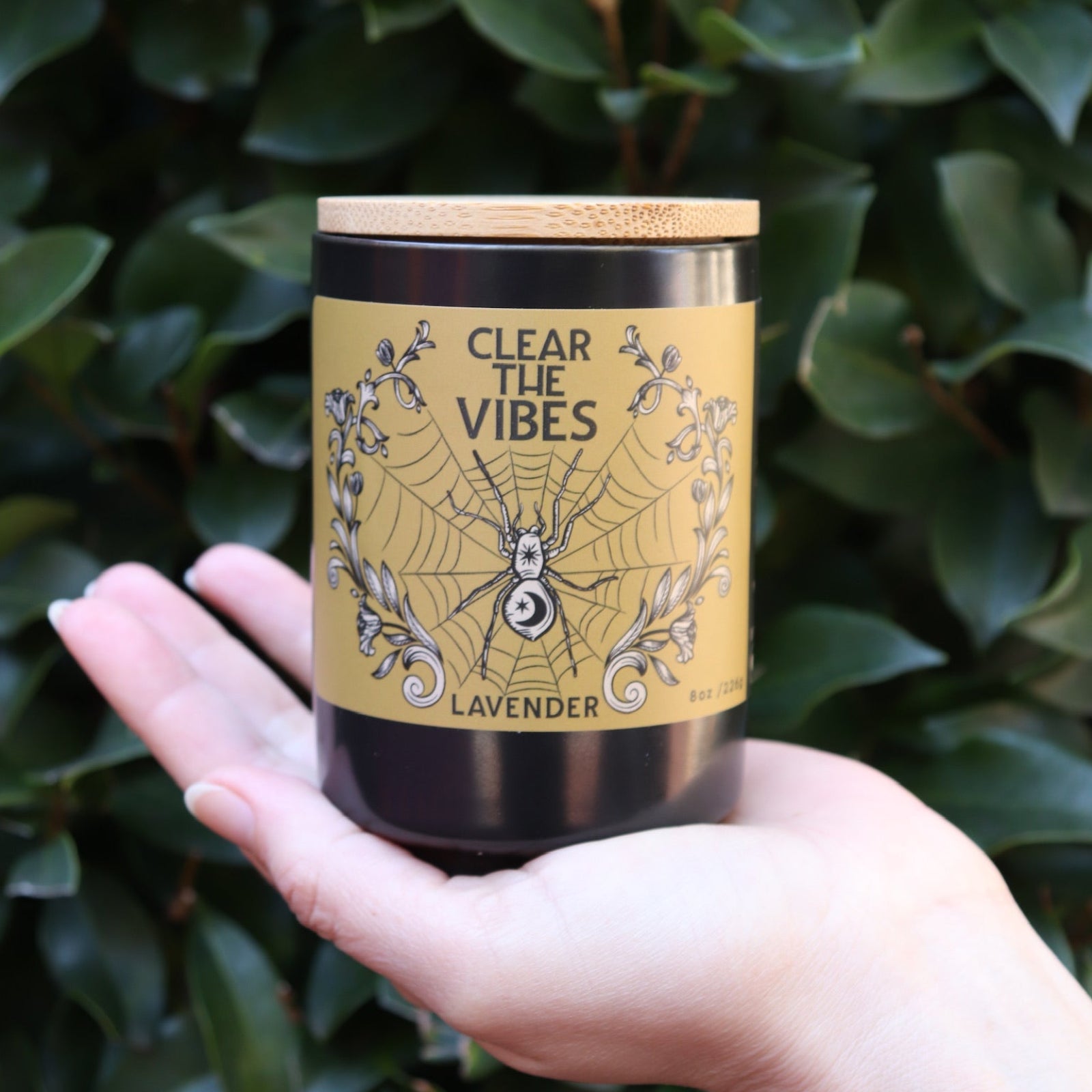 Someone holding the Clear the Vibes Soy Wax Garden Energy Clearing Candle in front of a green bush.