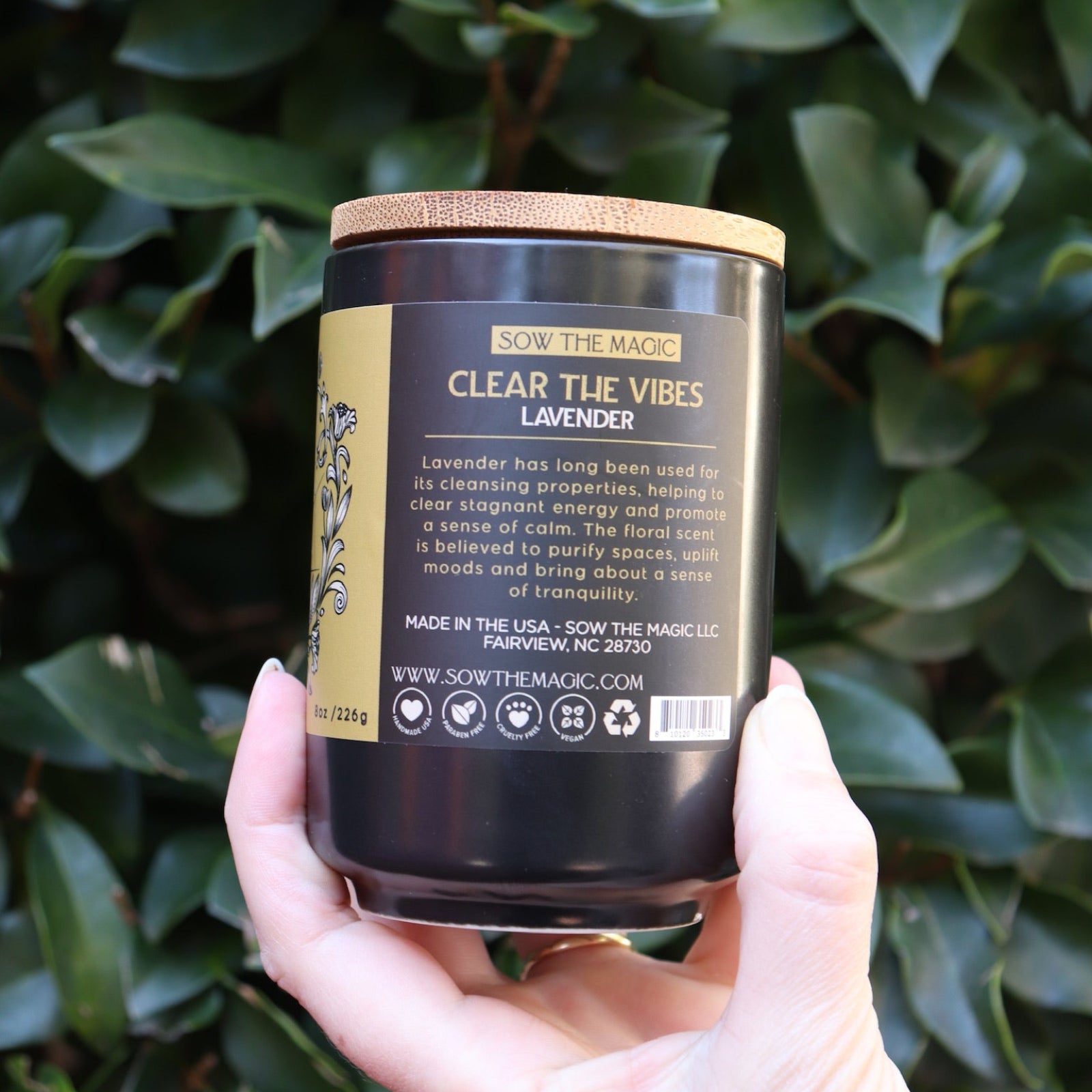 Someone holding the Clear the Vibes Soy Wax Garden Energy Clearing Candle in front of a green bush.