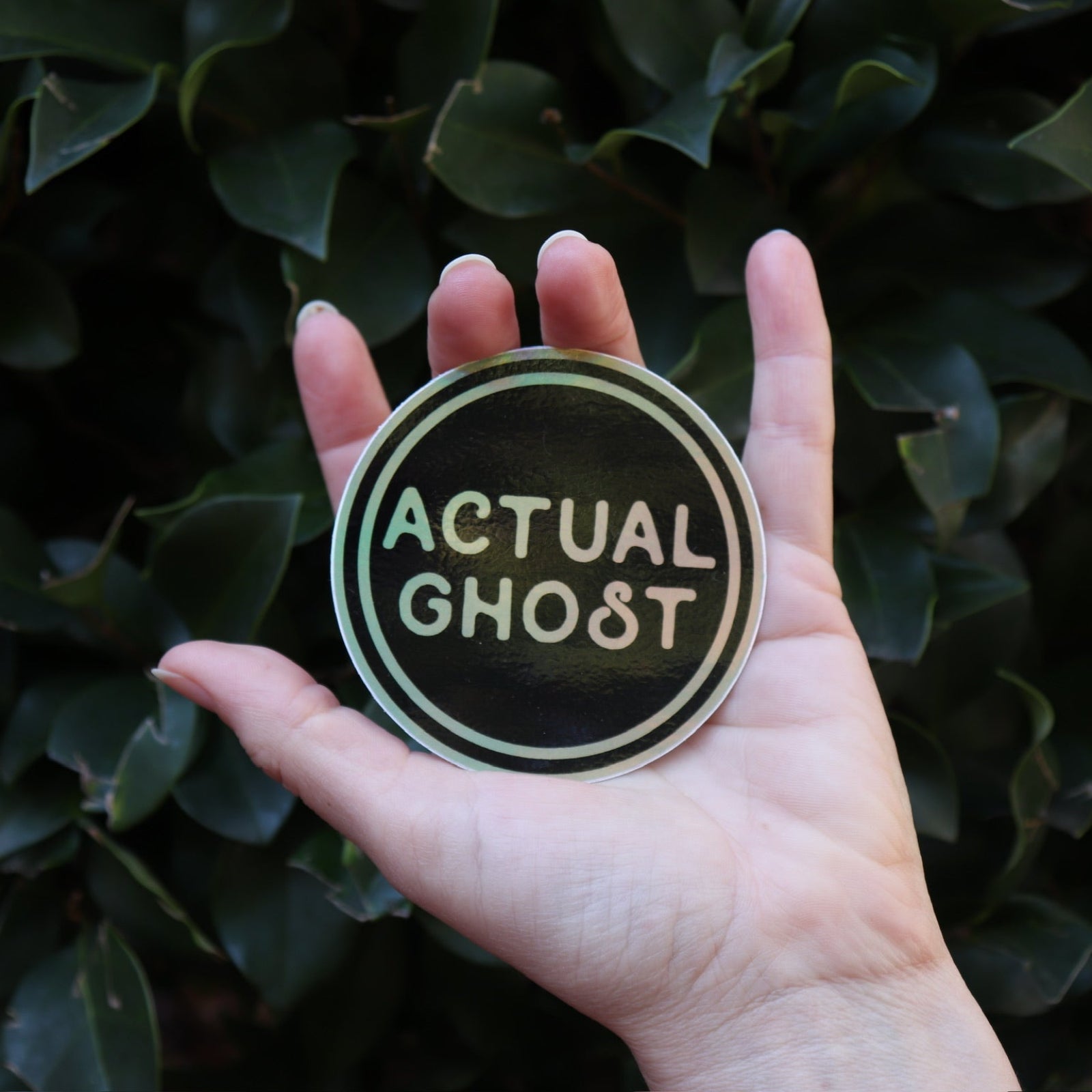 Someone holding the Actual Ghost Holographic Sticker in front of a green bush.