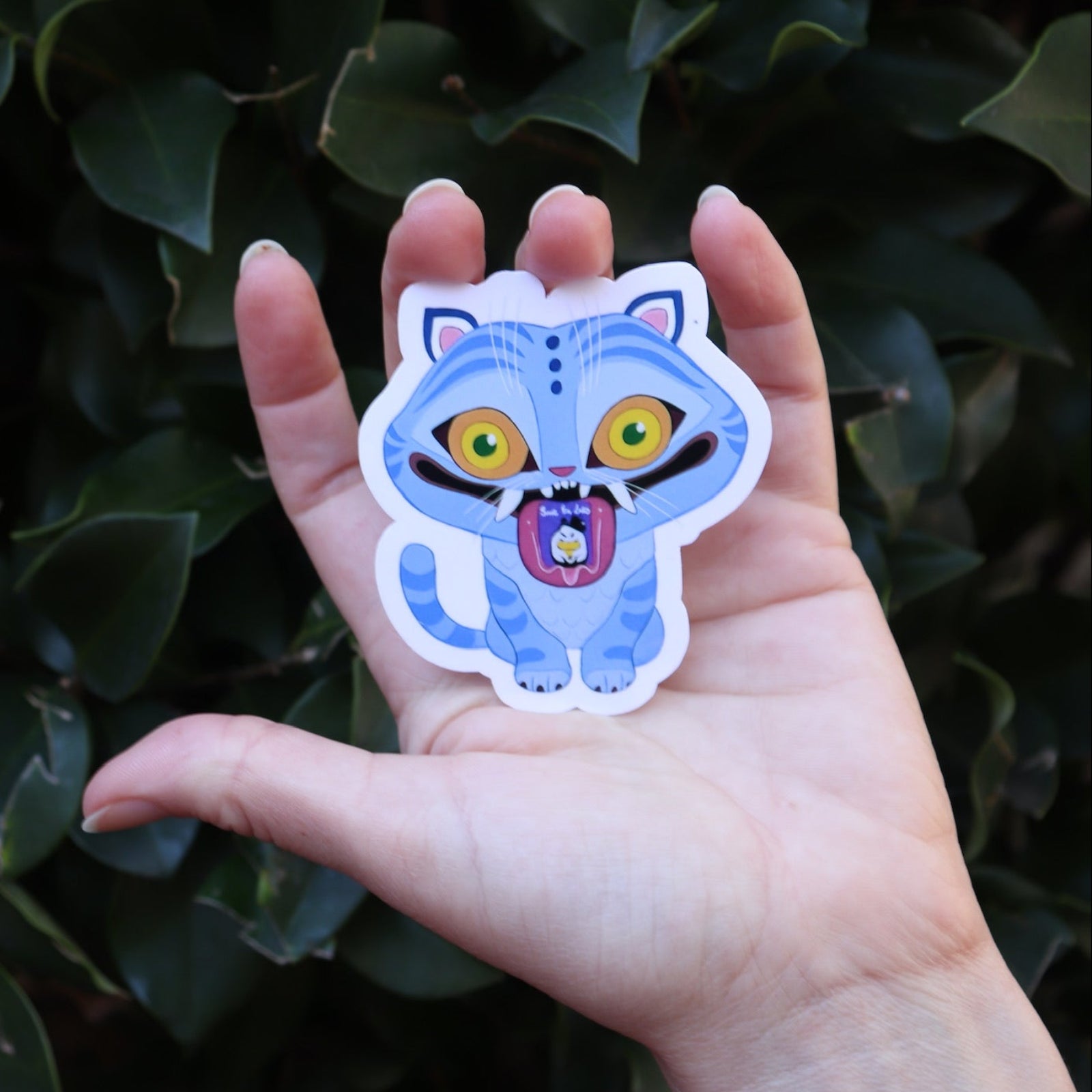 Someone holding the Derpy Tiger Kpop Demon Hunter Inspired Sticker in front of a green bush.