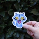 Someone holding the Derpy Tiger Kpop Demon Hunter Inspired Sticker in front of a green bush.