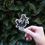 Someone holding the Sussie the Magpie KPop Demon Hunter Inspired Sticker in front of a green bush.