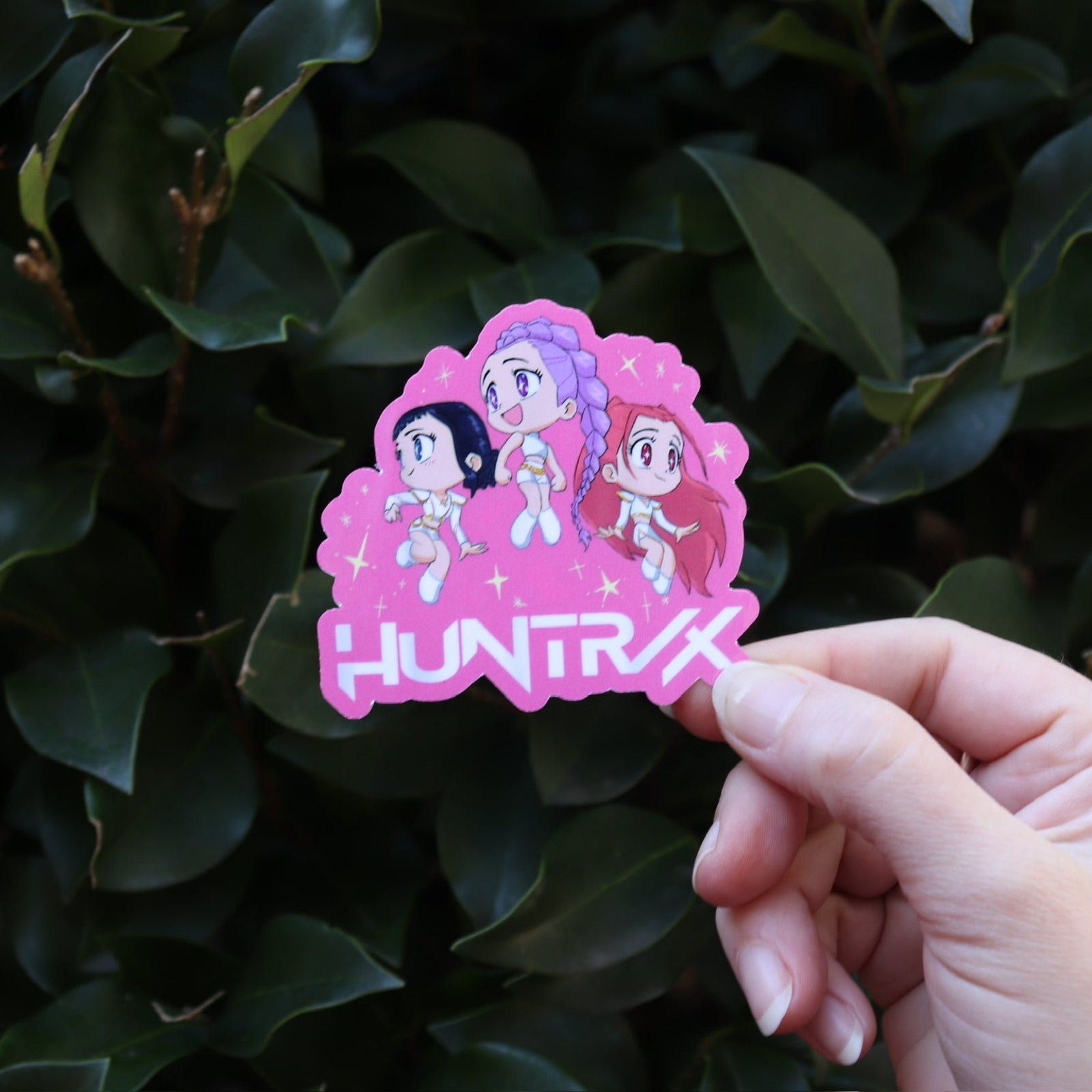 Someone holding the Chibi Huntrix KPop Demon Hunters Inspired Sticker in front of a green bush.