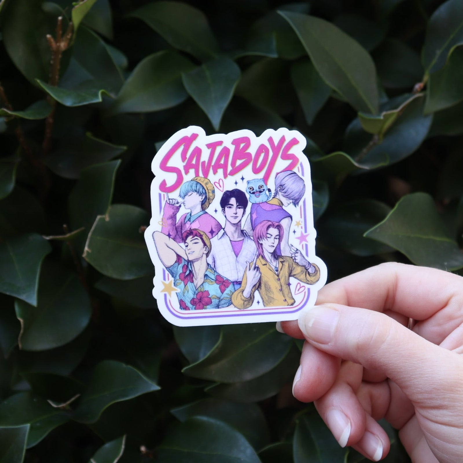 Someone holding the Saja Boys KPop Demon Hunters Inspired Sticker in front of a green bush.