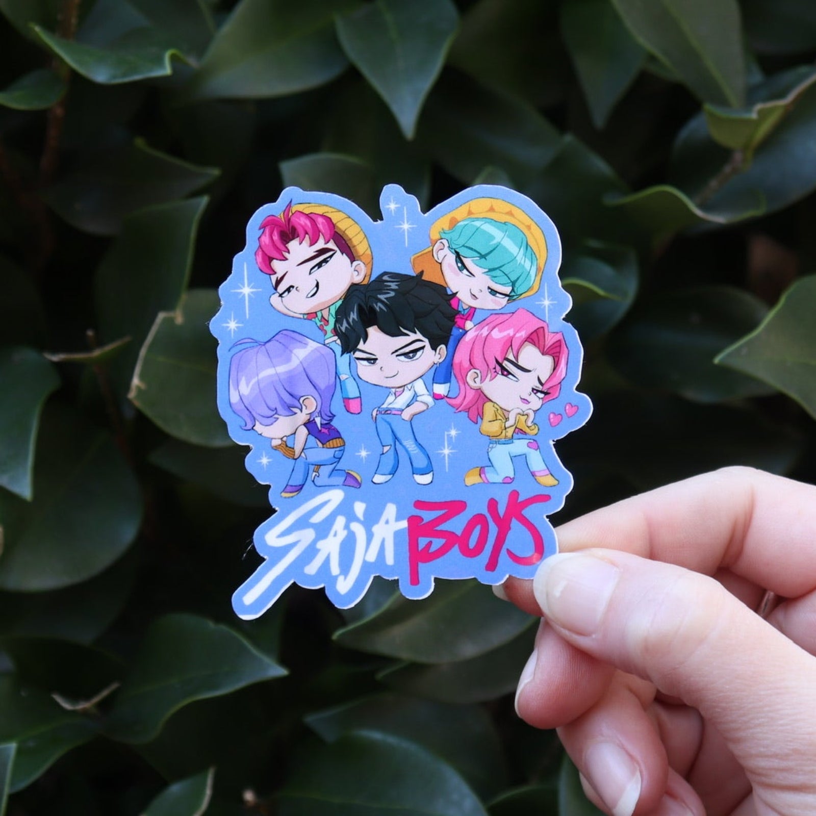 Someone holding the Chibi Saja Boys KPop Demon Hunters Inspired Sticker in front of a green bush.