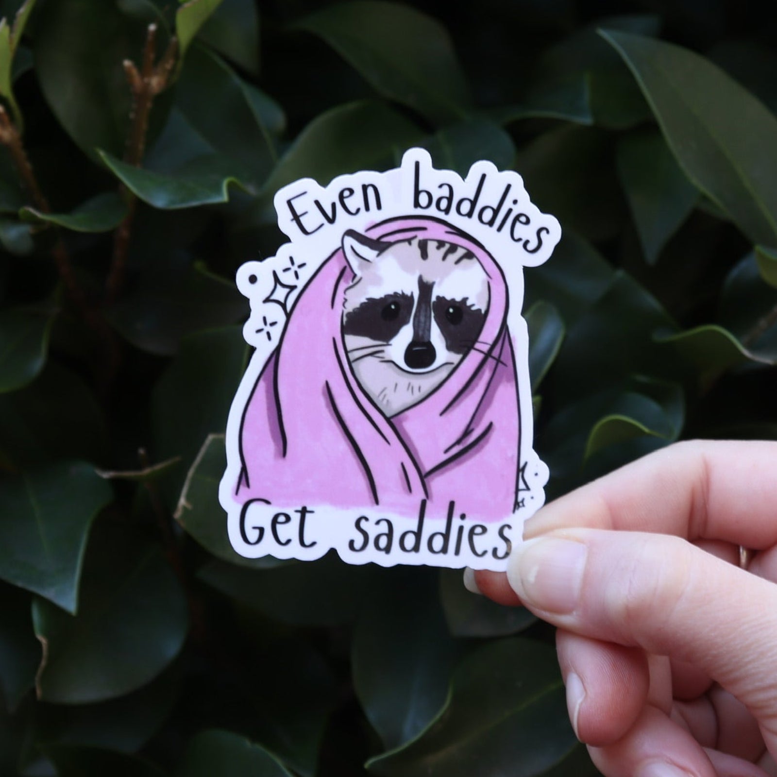 Someone holding the Even Baddies Get Saddies Funny Raccoon Sticker in front of a green bush.