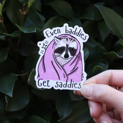 Someone holding the Even Baddies Get Saddies Funny Raccoon Sticker in front of a green bush.