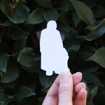 Someone holding the Slutty Little Knee Pedro Pascal Funny Sticker in front of a green bush, showing off the back.