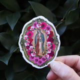 Someone holding the Virgen de Guadalupe Sticker in front of a green bush.