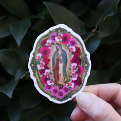 Someone holding the Virgen de Guadalupe Sticker in front of a green bush.