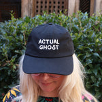 A woman wearing the Actual Ghost Black Unisex Baseball Cap in front of a green bush.