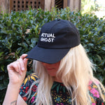 A woman wearing the Actual Ghost Black Unisex Baseball Cap in front of a green bush, showing the side.