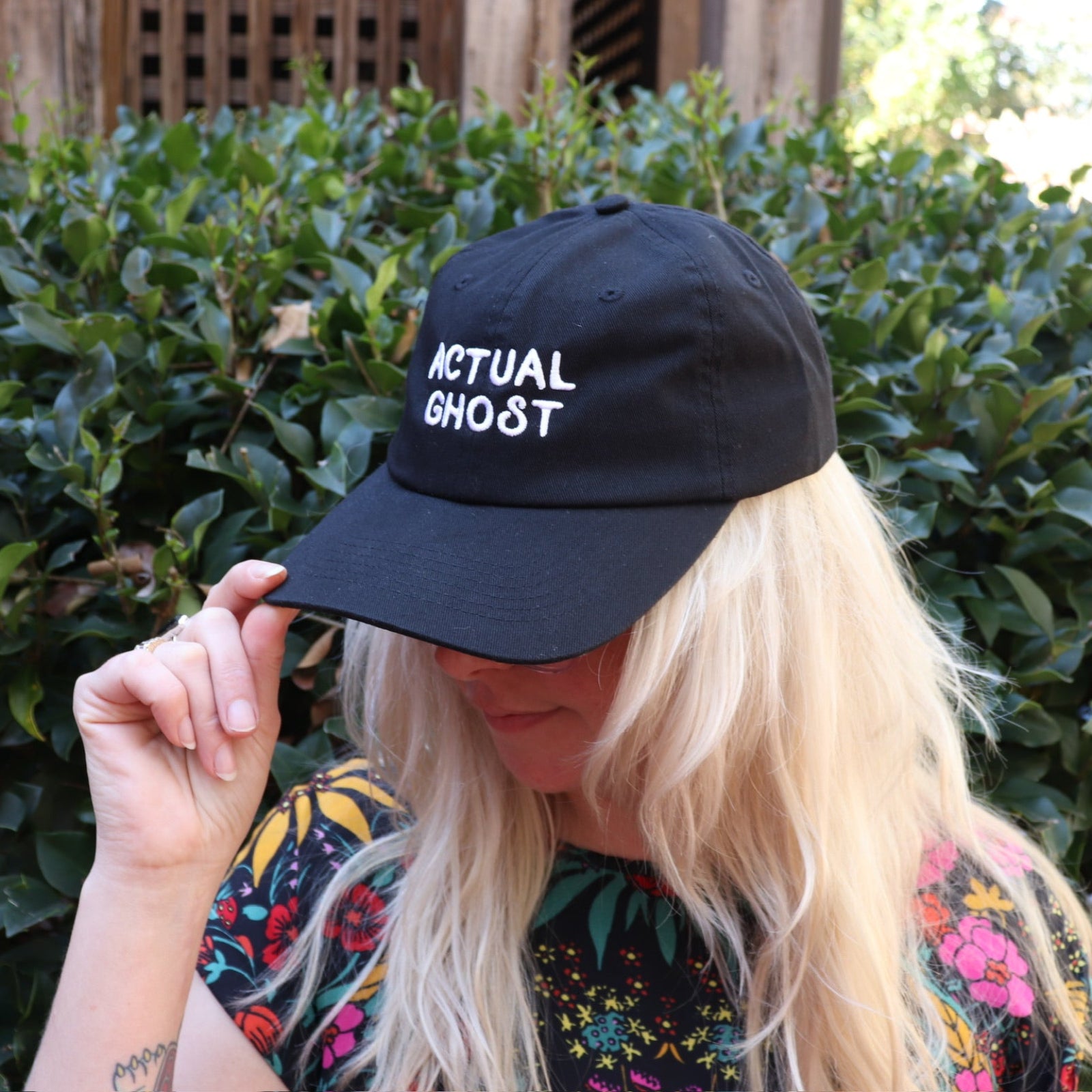 A woman wearing the Actual Ghost Black Unisex Baseball Cap in front of a green bush, showing the side.