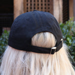 A woman wearing the Actual Ghost Black Unisex Baseball Cap in front of a green bush, showing off the back.