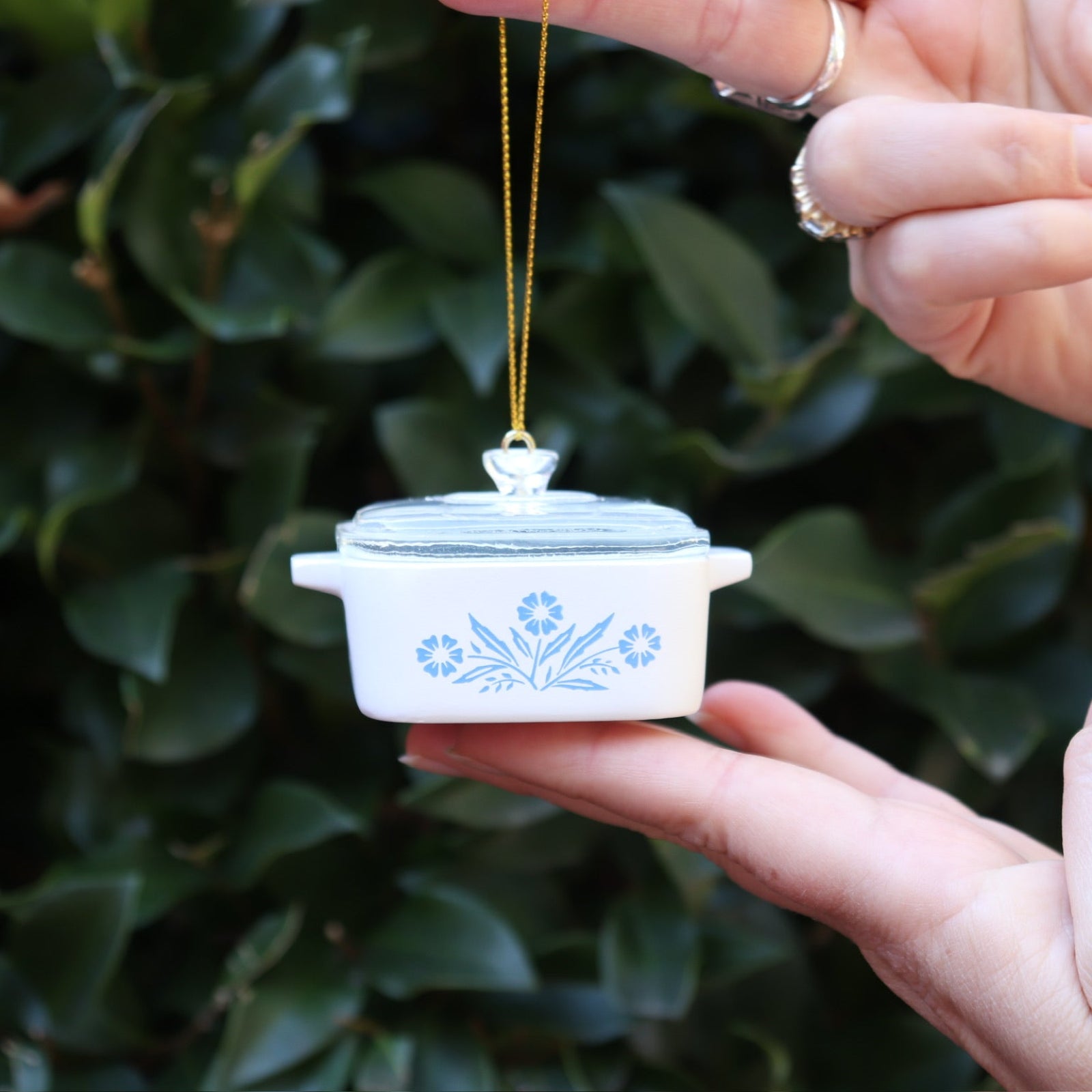 Someone holding the Mini Cornflower Casserole Ornament in front of a green bush.