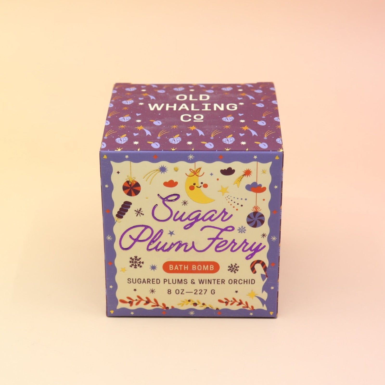 The Sugar Plum Ferry Holiday Bath Bomb in front of its box, all on a pink and yellow background.