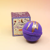 The Sugar Plum Ferry Holiday Bath Bomb in front of its box, all on a pink and yellow background.