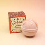 The Yuletide Glow Holiday Bath Bomb in front of its box all in front of a pink and yellow background.