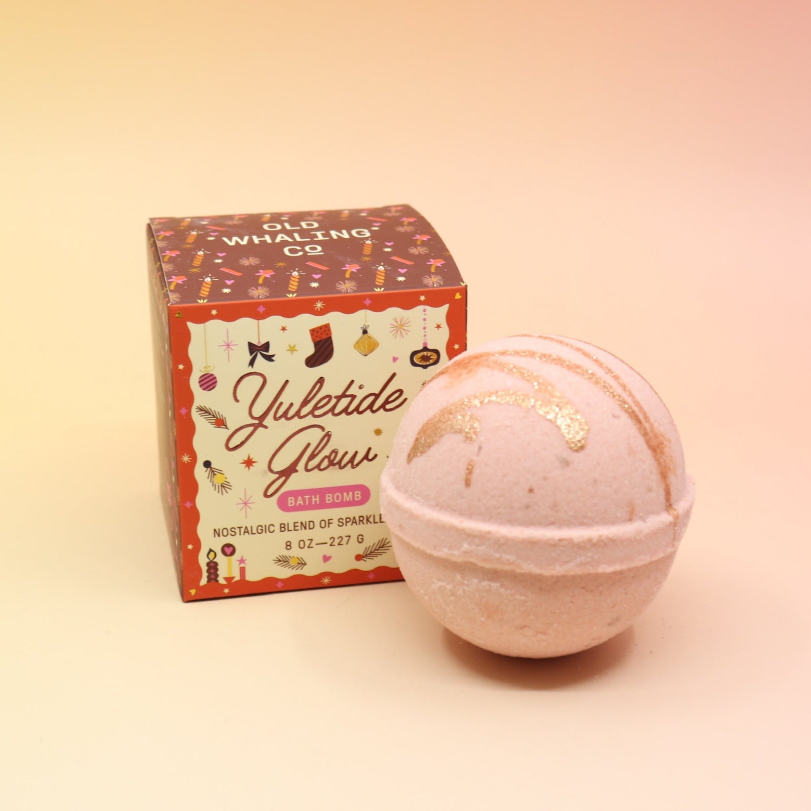 The Yuletide Glow Holiday Bath Bomb in front of its box all in front of a pink and yellow background.
