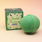 The Coastal Christmas Holiday Bath Bomb in front of its box all on a pink and yellow background.