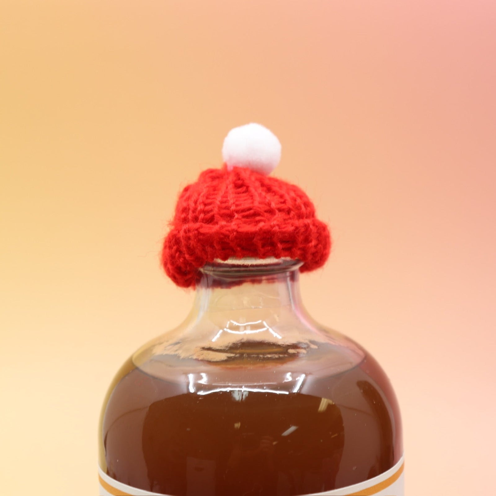 The red Mini Knit Cap on top of a bottle on a pink and yellow background.