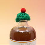 The green Mini Knit Cap on top of a bottle on a pink and yellow background.