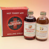The Winter Survival Gift Set with the two glass bottles in front of the box all on a pink and yellow background.