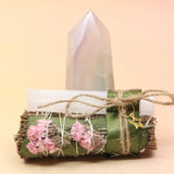 The Selenite Thyme Cleansing Smudge Bundle in front of a crystal tower on a pink and yellow background.