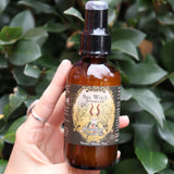 Someone holding the Krampus Scented Veil Spray in front of a green bush.