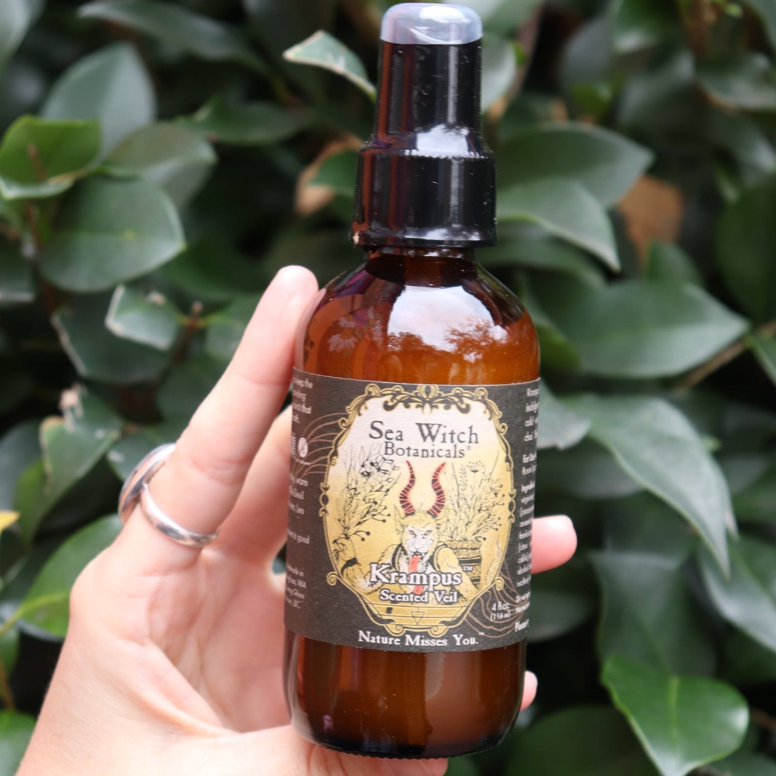 Someone holding the Krampus Scented Veil Spray in front of a green bush.