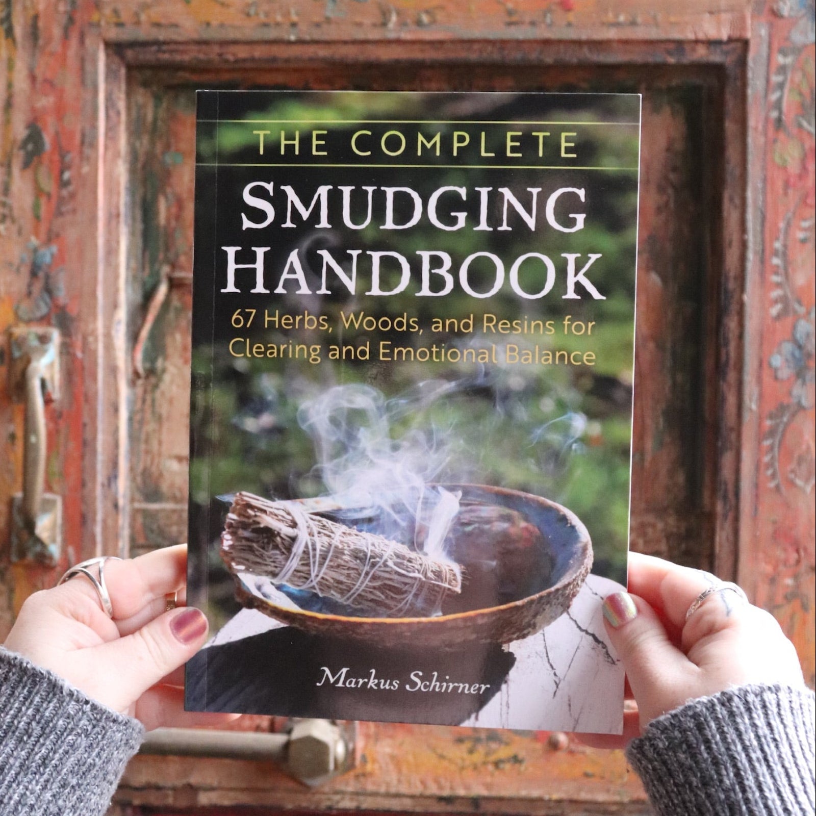 Someone holding the Complete Smudging Handbook Paperback Book in front of a wooden background.