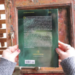 Someone holding the Complete Smudging Handbook Paperback Book in front of a wooden background, showing off the back.