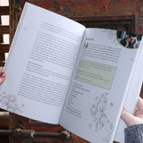 Someone holding the Complete Smudging Handbook Paperback Book open in front of a wooden background, showing off the pages.