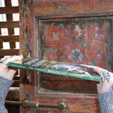 Someone holding the Complete Smudging Handbook Paperback Book in front of a wooden background, showing off the spine.