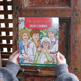 Someone holding the Golden Girls Color-by-Number Coloring Book in front of a wooden background.