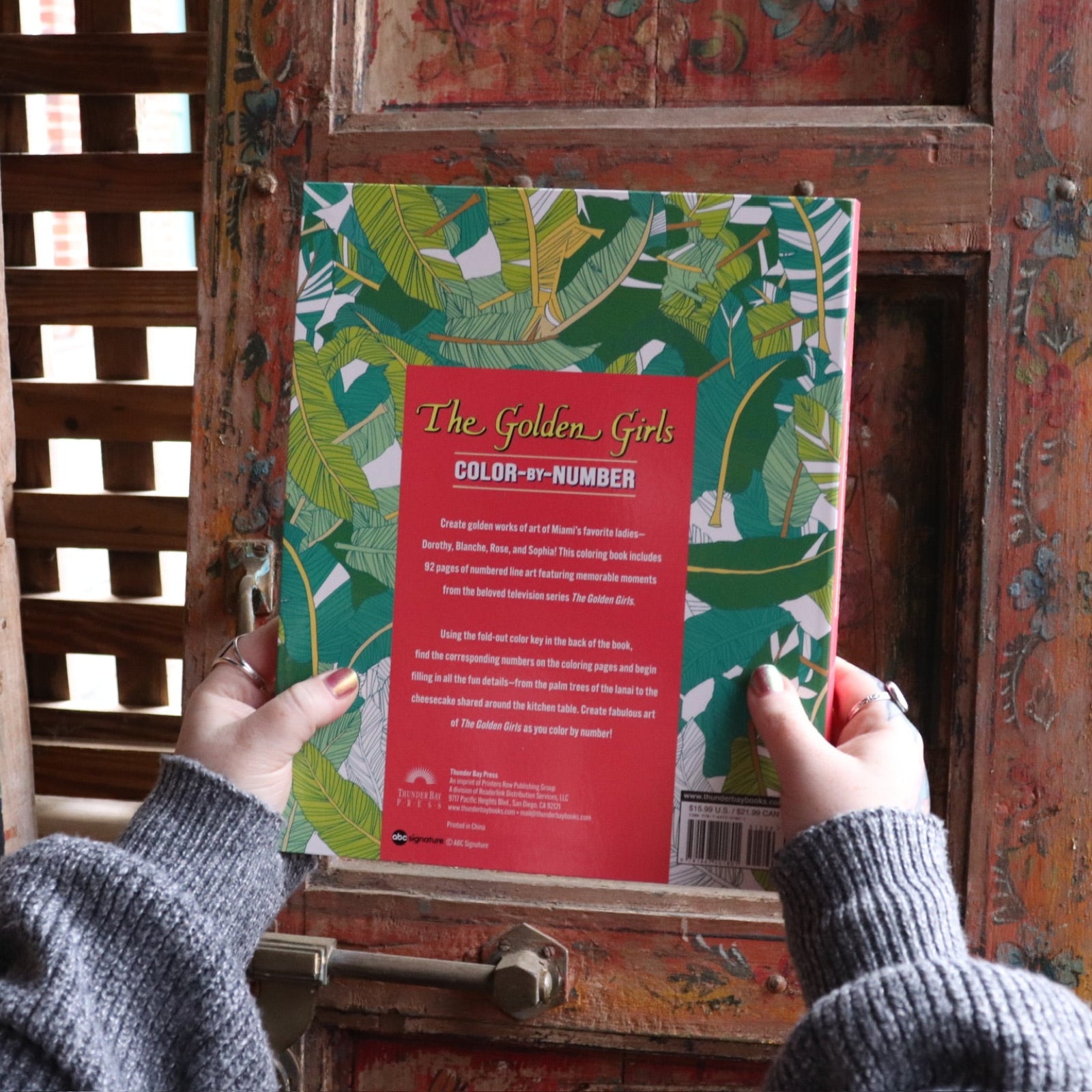 Someone holding the Golden Girls Color-by-Number Coloring Book in front of a wooden background, showing the background.