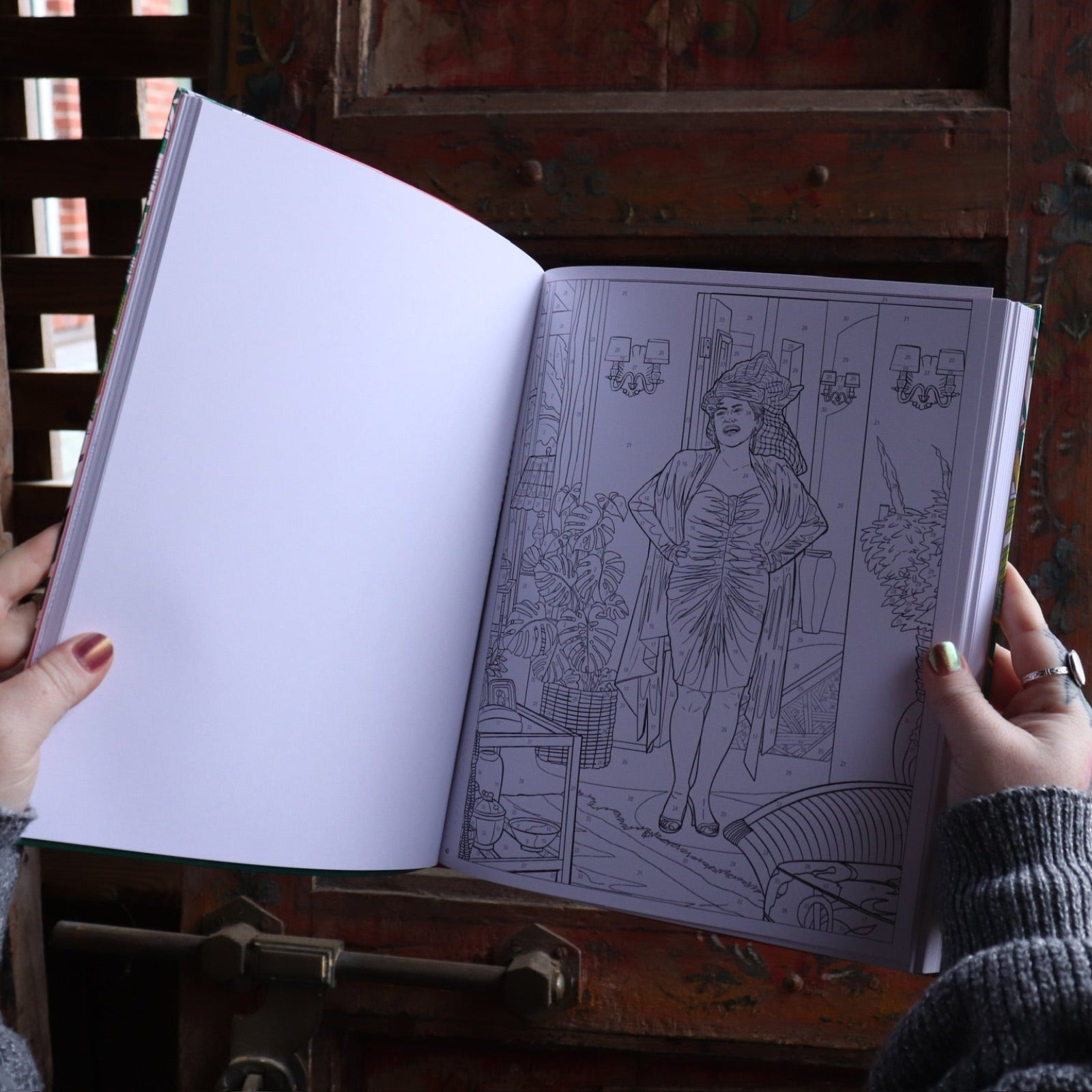 Someone holding the Golden Girls Color-by-Number Coloring Book open in front of a wooden background, showing off the pages.