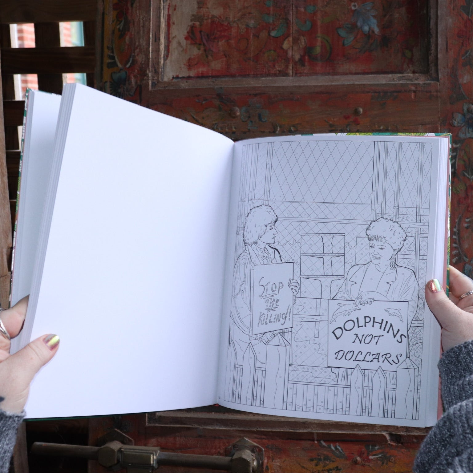 Someone holding the Golden Girls Color-by-Number Coloring Book open in front of a wooden background, showing off the pages.