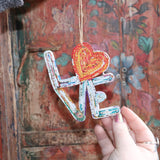 Someone holding the Recycled Magazine Love Heart Ornament in front of a colorful wooden background.