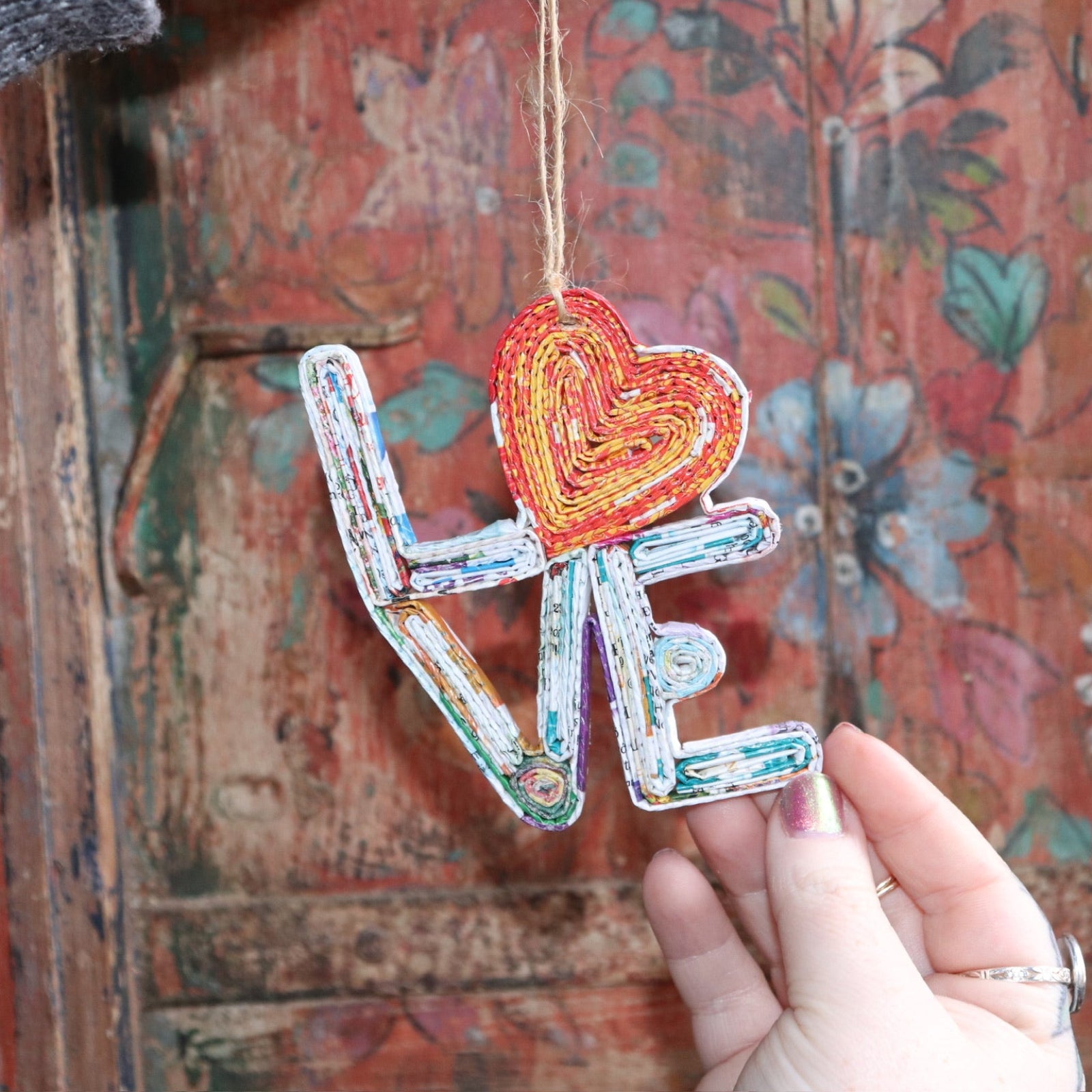 Someone holding the Recycled Magazine Love Heart Ornament in front of a colorful wooden background.