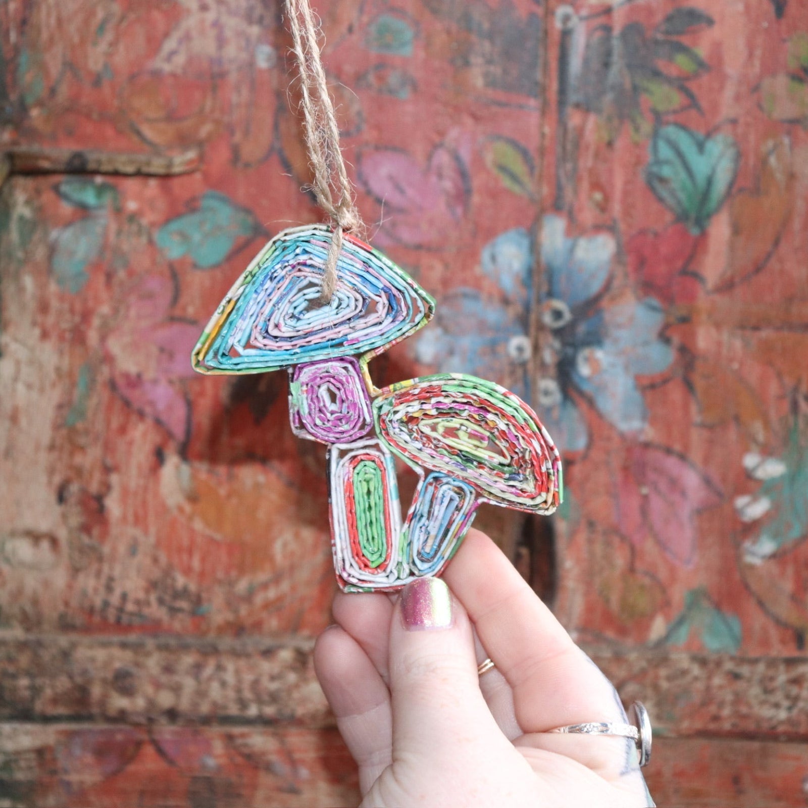 Someone holding the Recycled Magazine Mushroom Ornament in front of a colorful wooden background.