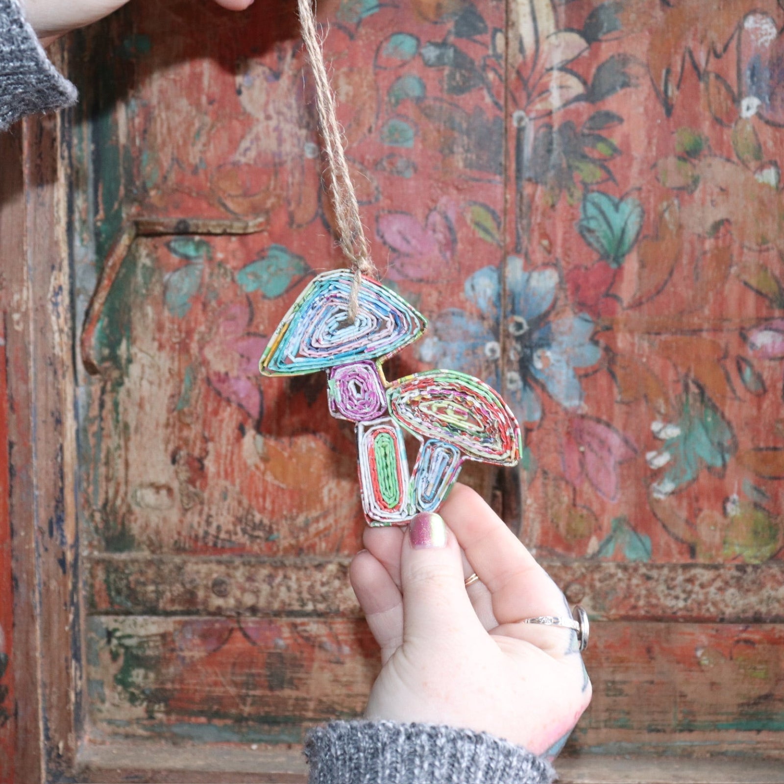 Someone holding the Recycled Magazine Mushroom Ornament in front of a colorful wooden background.