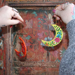 Someone holding two Papier-mâché Hand-Painted Moon Ornaments in front of a colorful wooden background.