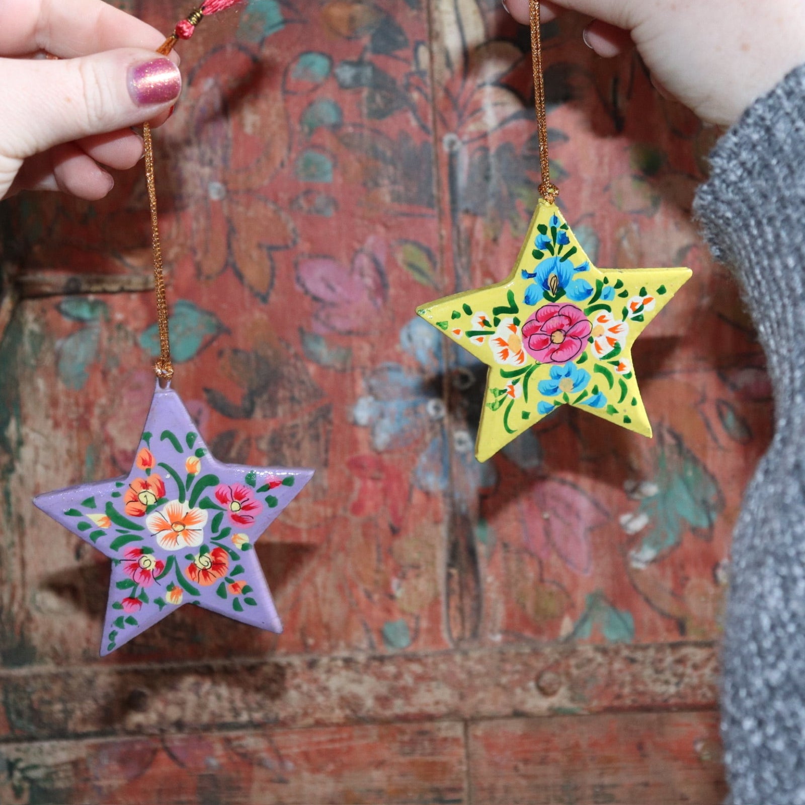 Someone holding two Papier-mâché Hand-Painted Star Ornaments in front of a colorful wooden background.