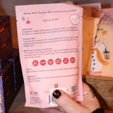 Someone holding a Pink Christmas Tree Bath Soak in front of other bath holiday products, showing off the back.